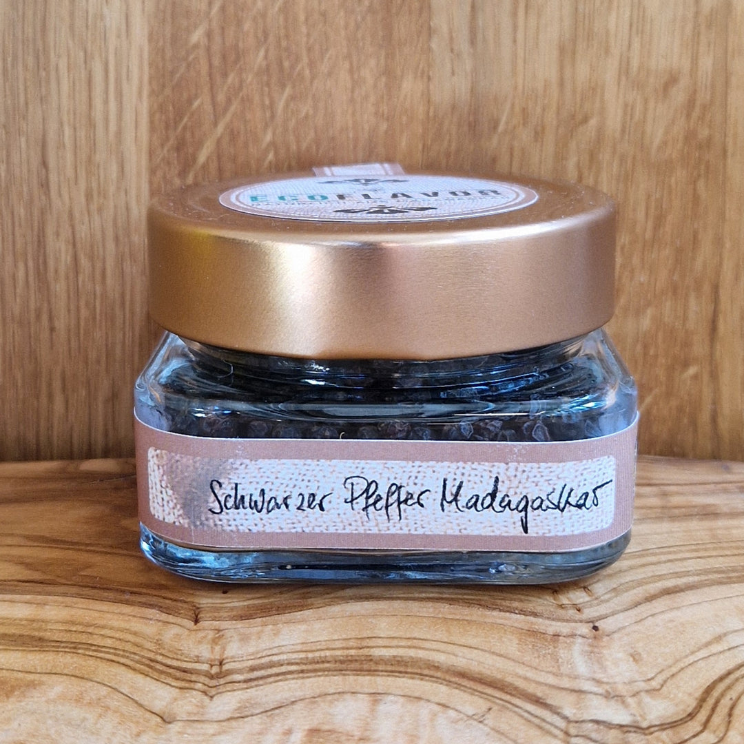 Schwarzer Pfeffer aus Madagaskar – naturbelassen, sonnengetrocknet und handverlesen, in einem Glas mit kupferfarbenen Deckel auf einem Holzbrett präsentiert.